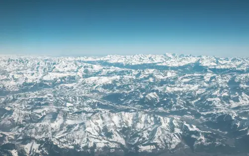 Aerial view of a vast mountain range covered in snow, with countless peaks stretching into the distance under a clear blue sky.