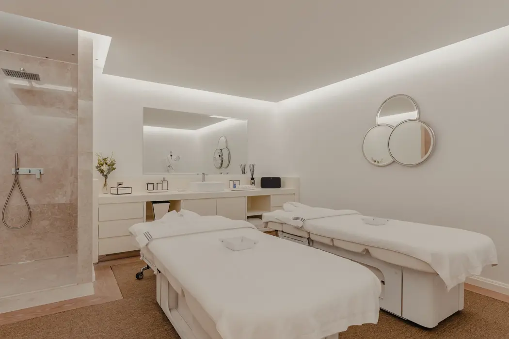 Luxury spa treatment room with two massage beds, minimalist décor, soft lighting, and a modern walk-in shower.