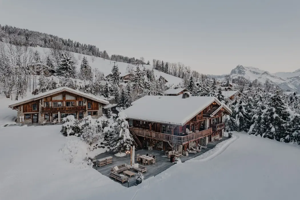 Luxury wooden chalets surrounded by snow-covered trees and mountains, with a spacious terrace and outdoor seating under a pale winter sky.