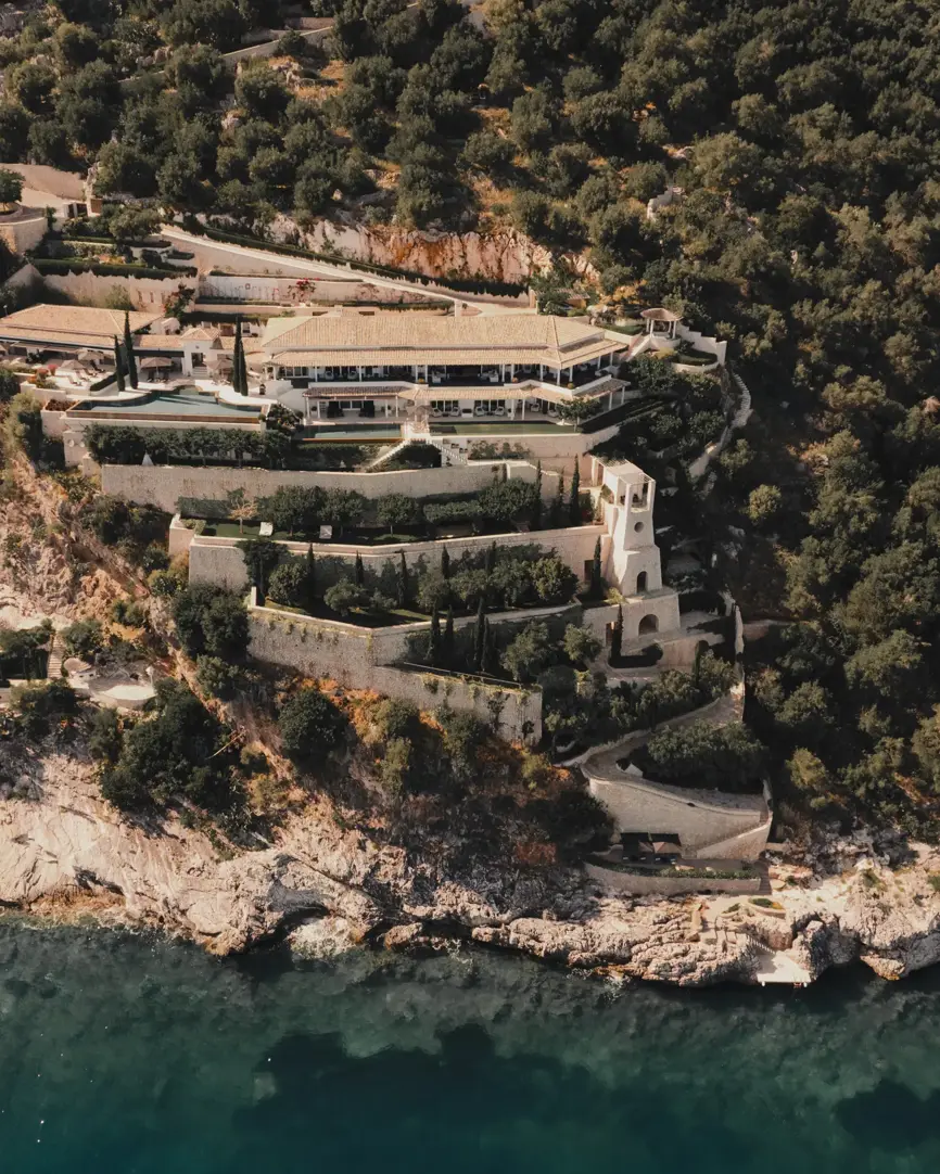 Aerial view of a luxury coastal villa built into a hillside with terraces, gardens, and a pool overlooking the turquoise sea.
