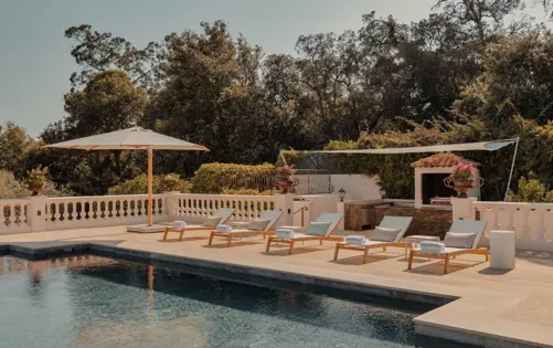 Elegant Mediterranean poolside terrace with sun loungers, white umbrellas, and outdoor kitchen surrounded by lush greenery under a clear sky.