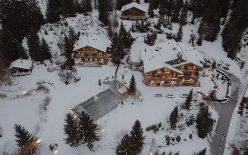 Aerial view of an exclusive alpine chalet estate surrounded by snow-covered pine forests, showcasing elegant wooden architecture, warm exterior lighting, and a heated outdoor pool in a private mountain resort.