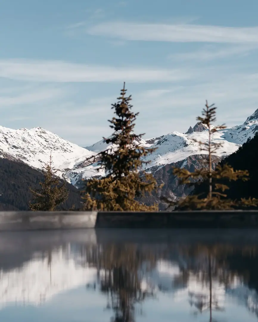 Snow-covered alpine mountains reflected in a still outdoor pool surrounded by pine trees.