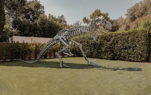 Polished metal Tyrannosaurus rex skeleton sculpture displayed on a manicured lawn surrounded by lush greenery and modern landscaping.