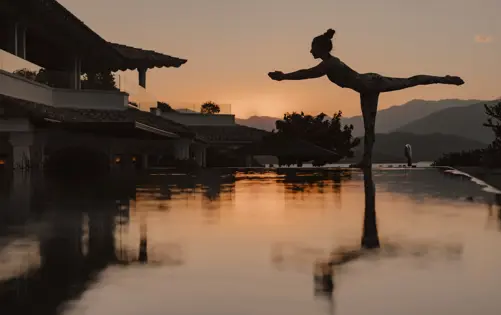 Silhouette of a woman practicing yoga in a balancing pose beside an infinity pool at sunset, with mountains and a tranquil sky in the background.