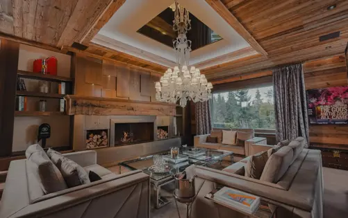 Elegant living room in a luxury alpine chalet featuring a grand crystal chandelier, modern fireplace, velvet sofas, and panoramic forest views. The refined mix of wood, glass, and designer accents creates an inviting yet sophisticated mountain retreat.