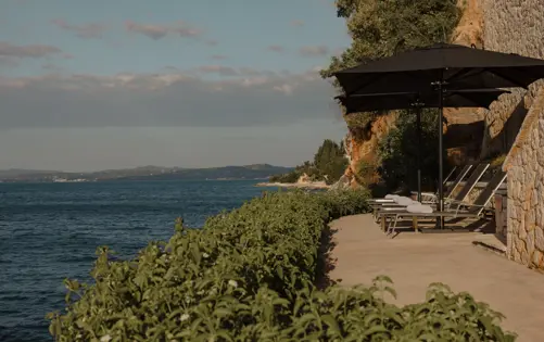 Seaside terrace built into a rocky cliff, featuring sun loungers with rolled white towels under black umbrellas, overlooking the calm blue sea and distant coastline.