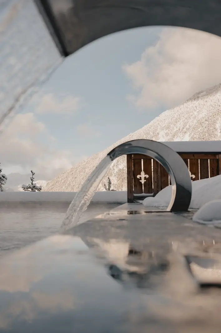 Outdoor heated pool with modern water features overlooking snow-covered mountains under a clear winter sky.