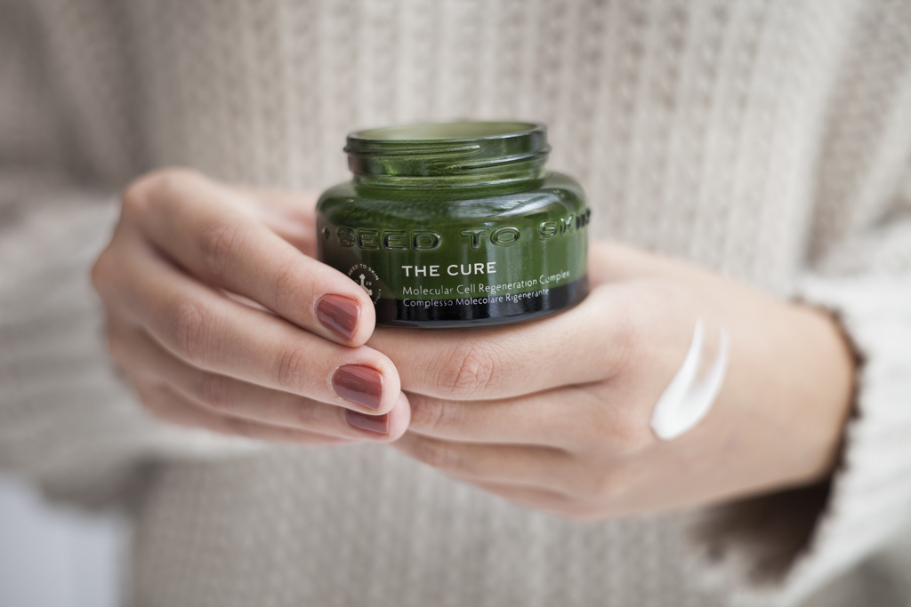 Hands holding a jar of Seed to Skin “The Cure” face cream with a small amount of cream applied to the skin.