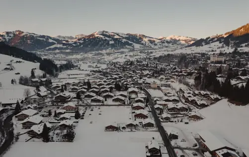 Aerial view of a snow-covered alpine village surrounded by mountains at sunrise, with wooden chalets and winding roads nestled in the valley.