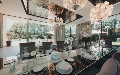 Luxurious dining room with a mirrored table, crystal chandeliers, and floor-to-ceiling windows opening to a sunlit terrace and garden view.