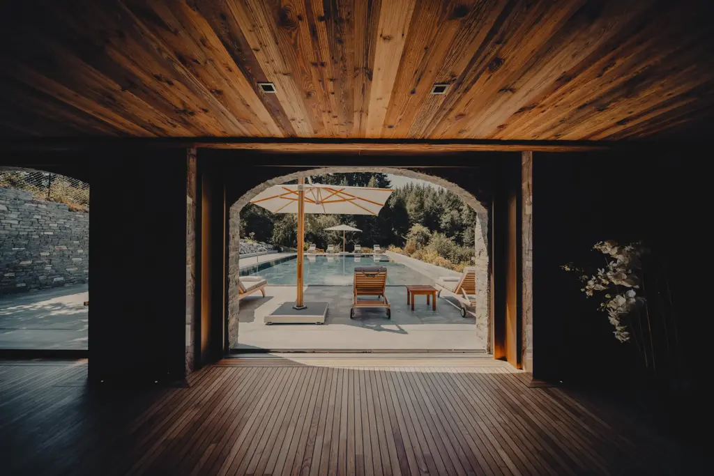 View from a wooden interior leading to an outdoor pool area with sun loungers, umbrellas, and lush greenery in the background.