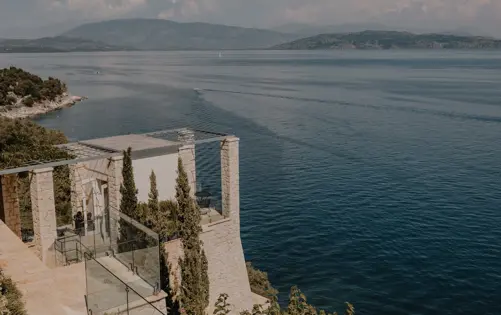 Elegant stone villa perched on a coastal cliff with glass terraces overlooking the tranquil blue sea and distant mountains.