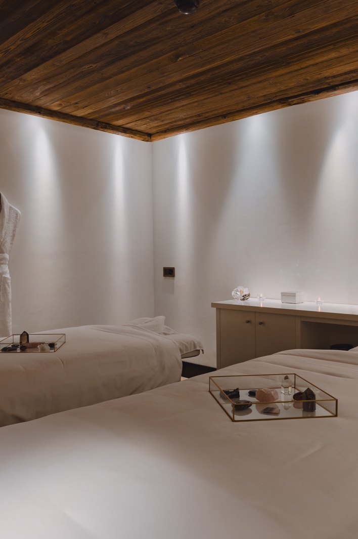 Luxurious spa room with two massage beds, soft lighting, wooden ceiling, and robes hanging on the wall.