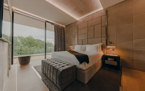 Luxurious modern bedroom with soft lighting, upholstered walls, large glass doors opening to a terrace, and a serene nature view.