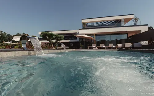 Modern luxury villa with an elegant infinity pool, cascading water features, sun loungers, and glass architecture under a clear blue sky.