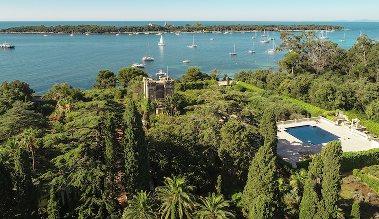 Aerial view of a lush Mediterranean estate with gardens, a historic villa, and a large pool overlooking the sea with sailboats and islands in the background.