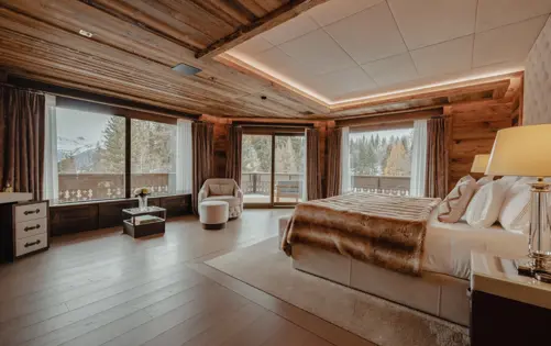 Spacious master bedroom in a luxury alpine chalet with panoramic forest and mountain views, natural wood paneling, soft ambient lighting, and a king bed dressed in fur and linen for an elegant mountain retreat.