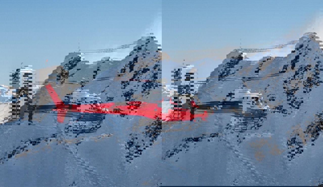 Red rescue helicopter flying over snowy Swiss Alps near a mountain station under a clear blue sky.