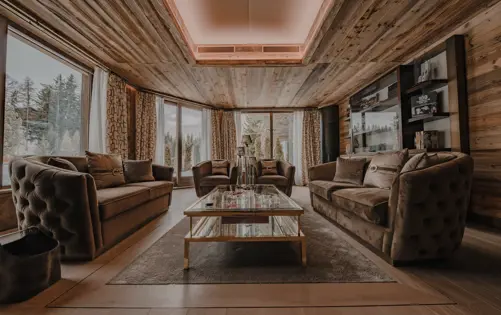 Elegant living room in a luxury alpine chalet featuring plush velvet sofas, a glass coffee table with gold accents, wood-paneled walls and ceiling, and panoramic forest views through large floor-to-ceiling windows.