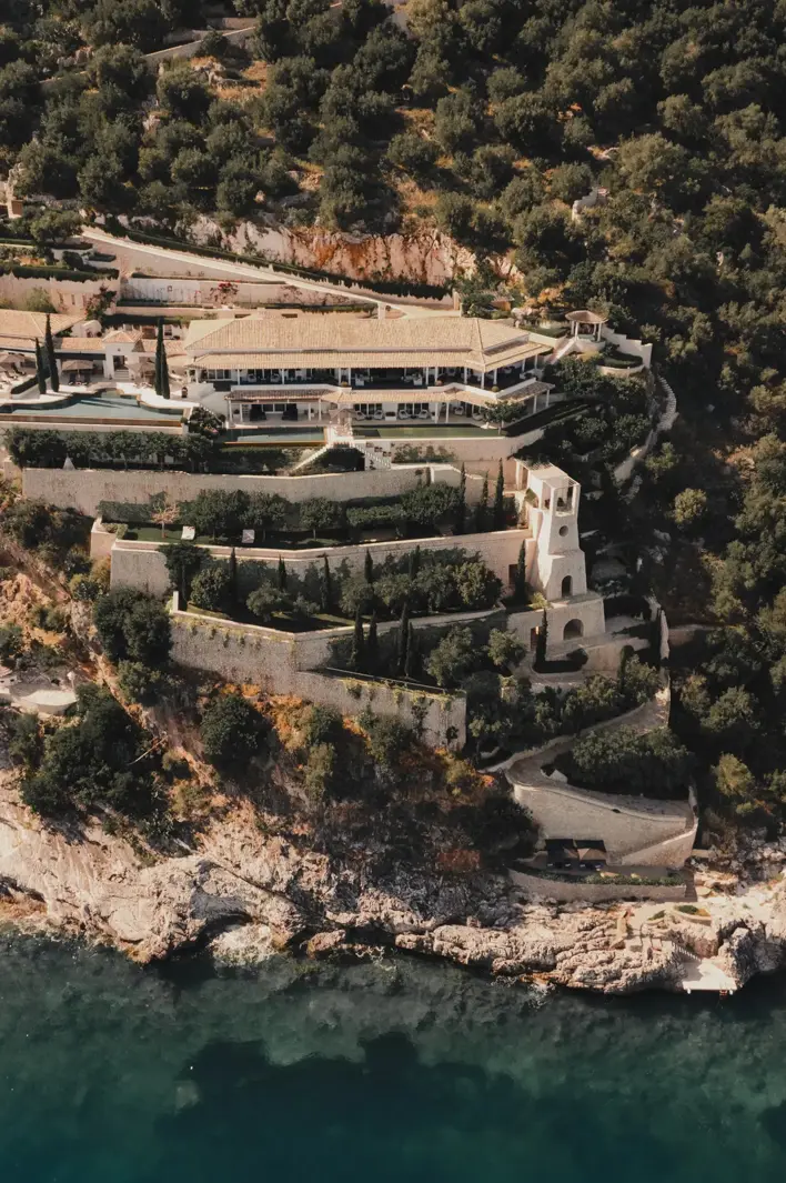 Vue aérienne d’un domaine méditerranéen de luxe perché sur une falaise, entouré de verdure luxuriante, de jardins en terrasses et dominant la mer turquoise.