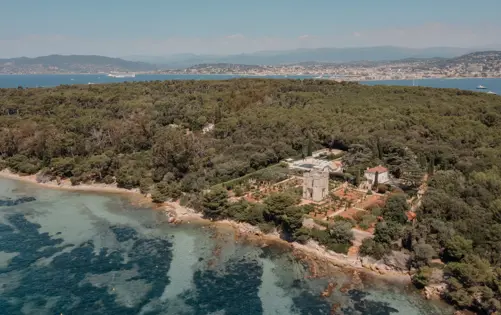 Aerial view of a historic estate surrounded by forest on a Mediterranean island, with turquoise water and a coastal town in the distance.