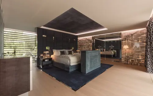 Contemporary luxury bedroom with modern lighting design, marble wall accents, sleek furnishings, and large windows overlooking greenery.