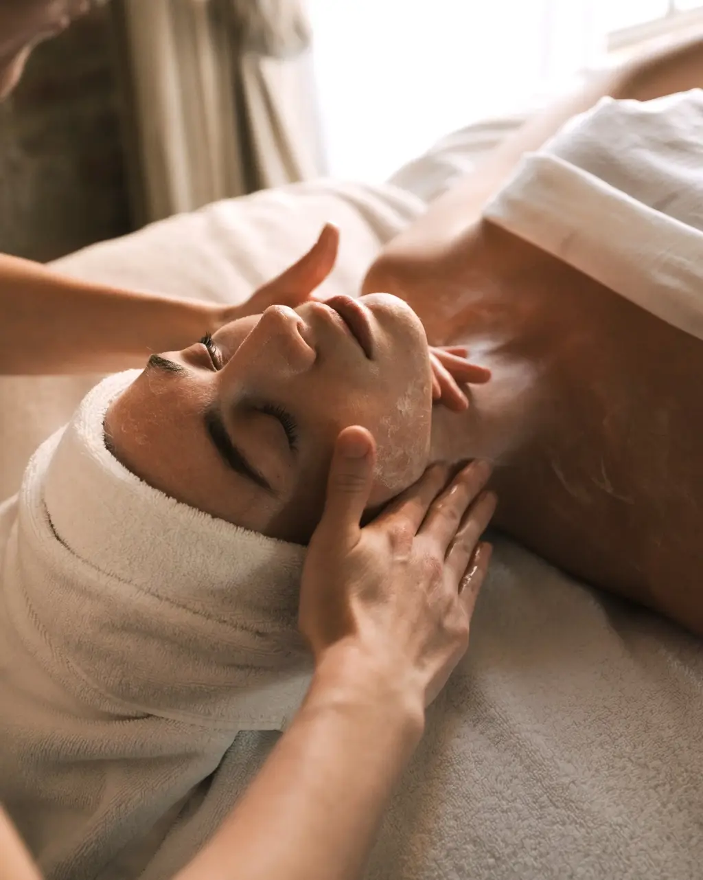 Spa therapist performing a relaxing facial massage on a person wrapped in white towels during a skincare treatment.