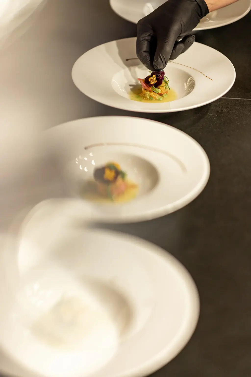 A chef wearing black gloves carefully places a garnish on an artfully plated dish, capturing the elegance and precision of haute cuisine.