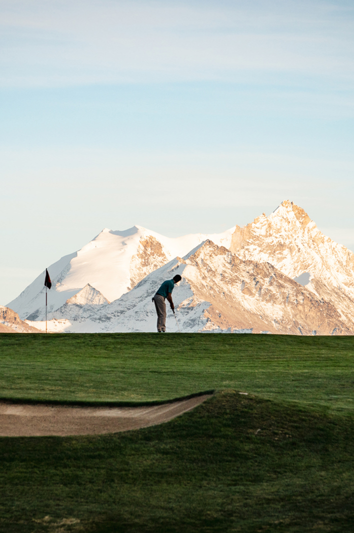 A golfer lines up a putt on a lush green course framed by majestic snow-capped mountains glowing in the golden morning light.