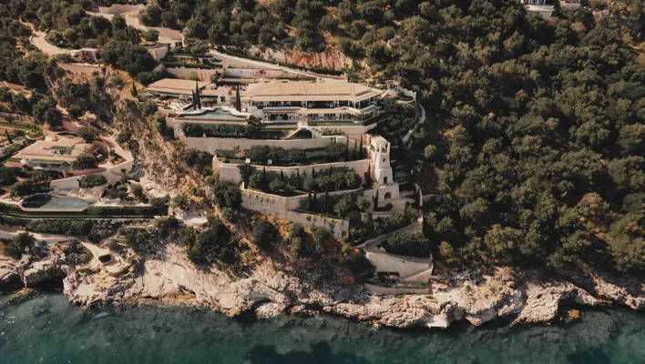 Aerial view of a luxury coastal villa built into a hillside with terraces, gardens, and a pool overlooking the turquoise sea.
