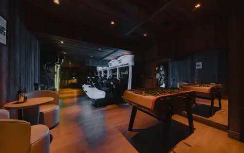 Luxury chalet game room with arcade racing simulators, foosball table, and lounge seating in a cozy wooden interior.