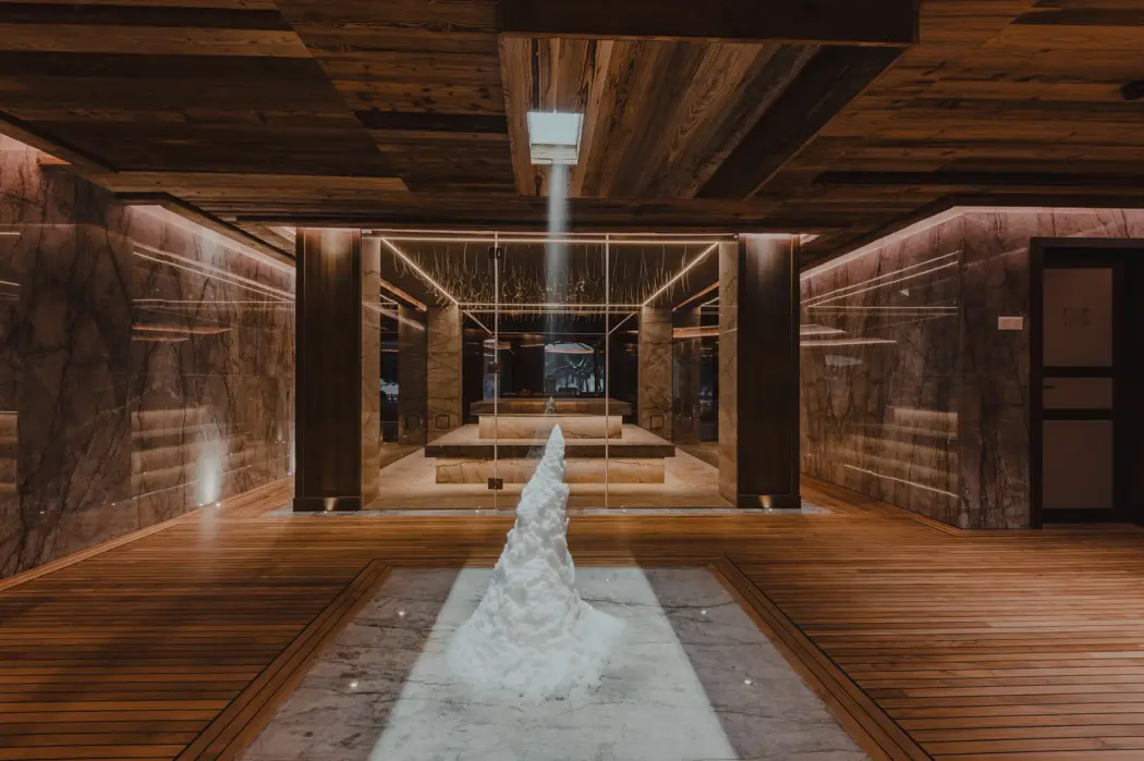 Contemporary snow room within a luxury alpine chalet spa, featuring a sculpted mound of real snow under a focused light beam, surrounded by marble walls and warm wooden finishes. Designed to enhance circulation and balance after sauna sessions, it offers a serene, sensory wellness experience inspired by Nordic traditions.