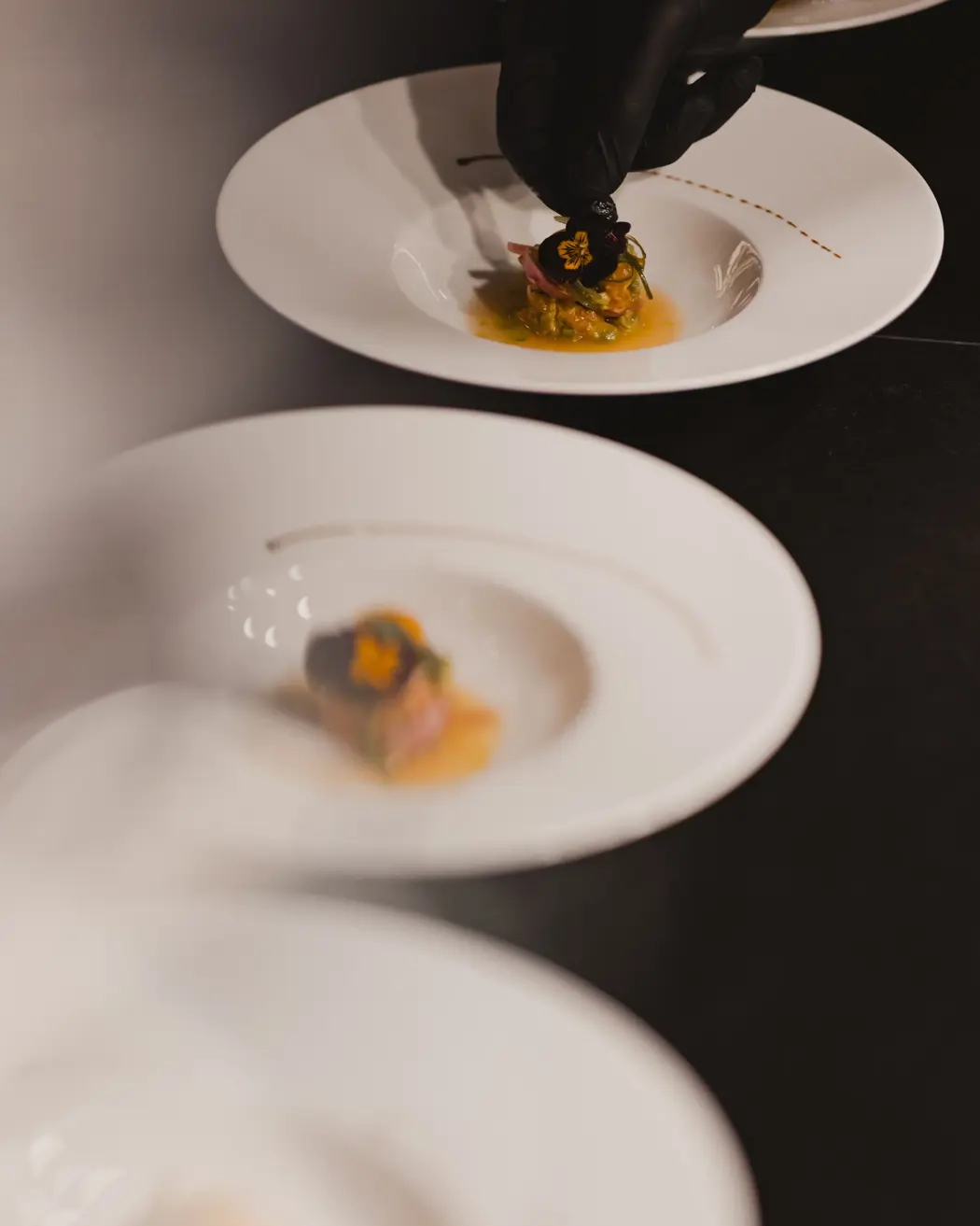 Chef’s hand plating a fine dining dish with precision, garnished with edible flowers on a white plate.