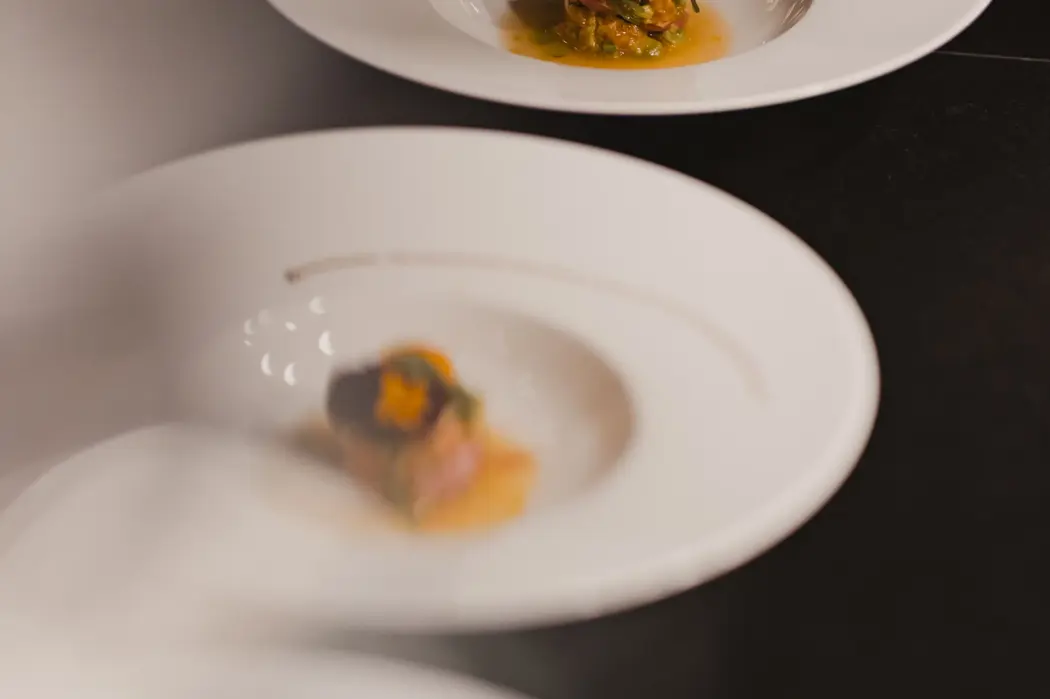Chef’s hand plating a fine dining dish with precision, garnished with edible flowers on a white plate.