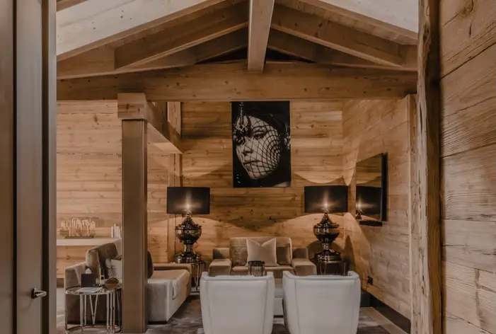 Уютная и элегантная гостиная в стиле шале со светлыми деревянными стенами и балками, мягкой мебелью, тёмными лампами и эффектным чёрно-белым портретом в центре композиции.