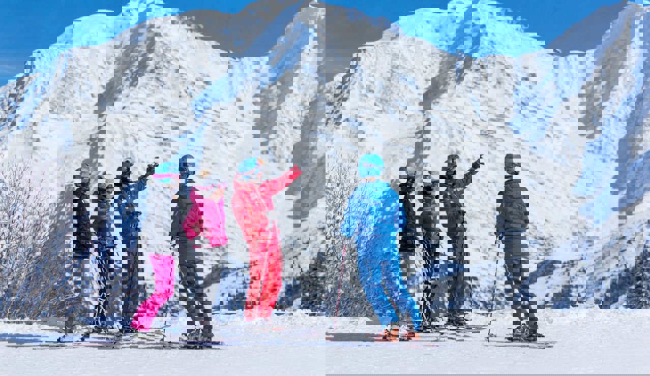 A ski instructor in a red outfit teaching three skiers on a snowy mountain slope, with tall snow-covered peaks in the background under a clear blue sky.