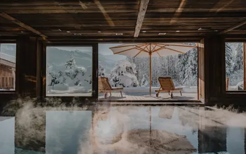Luxury chalet spa with indoor heated pool and panoramic mountain view, featuring sun loungers and umbrella on a snowy terrace.