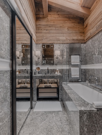 Роскошная ванная комната в стиле шале с элегантными серыми мраморными стенами и полом, ванной и умывальником под наклонным деревянным потолком с балками и мансардным окном, наполняющим пространство естественным светом.