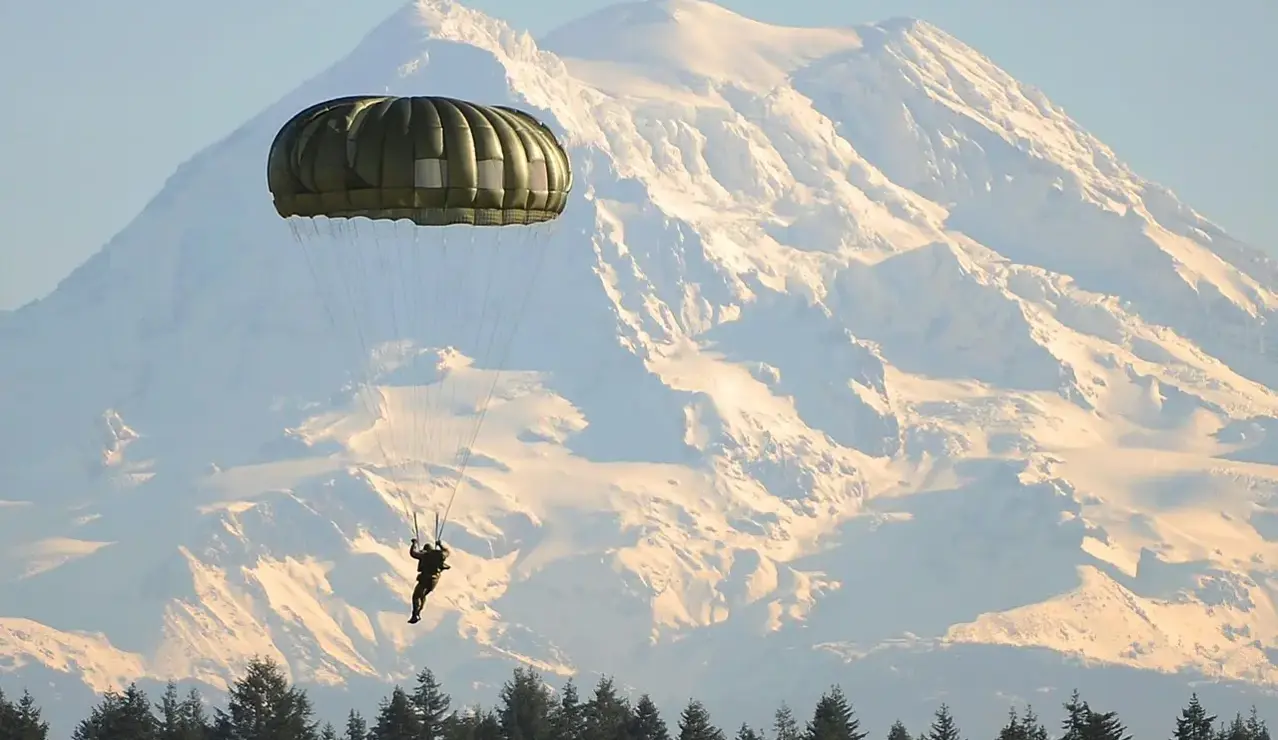 A lone parachutist descends through clear alpine air with snow-capped peaks rising in the distance — an image of freedom, precision, and mountain adventure.