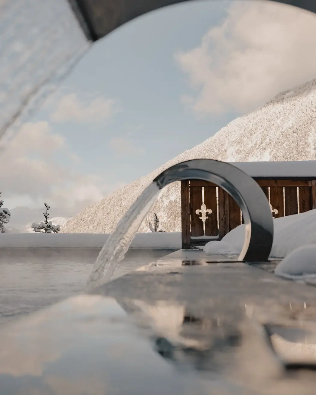 Outdoor heated pool with modern water features overlooking snow-covered mountains under a clear winter sky.