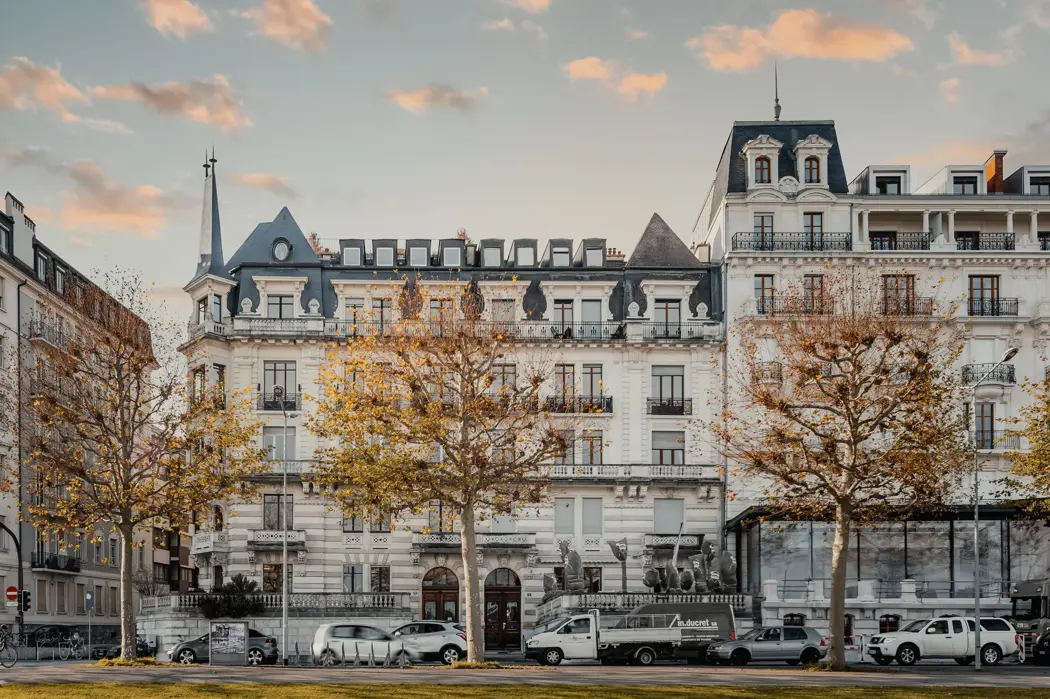 Elegant historic European building with ornate architecture, large windows, and mansard roofs, lined with autumn trees and parked cars under a pastel sunset sky.