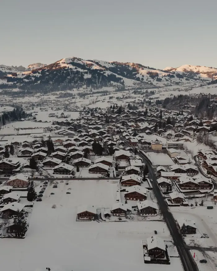 Aerial view of a snow-covered alpine village surrounded by mountains at sunrise, with wooden chalets and winding roads nestled in the valley.