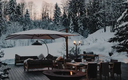 Elegant chalet terrace in winter with snow-covered forest, fire pit, dining table, and lounge seating under large parasol at sunset.