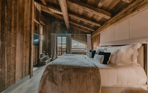 Luxury chalet master bedroom with wooden walls and ceiling, soft lighting, mountain view, and plush bedding in an alpine retreat.