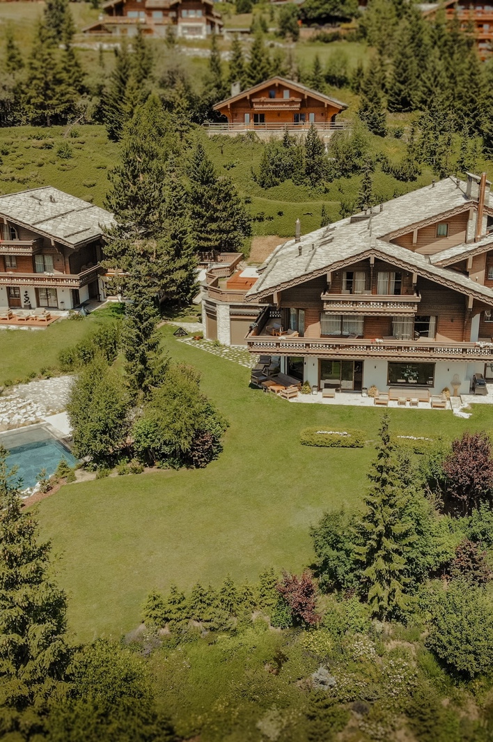 Aerial view of a luxury alpine chalet surrounded by pine trees with a pool, terraces, and mountain scenery.