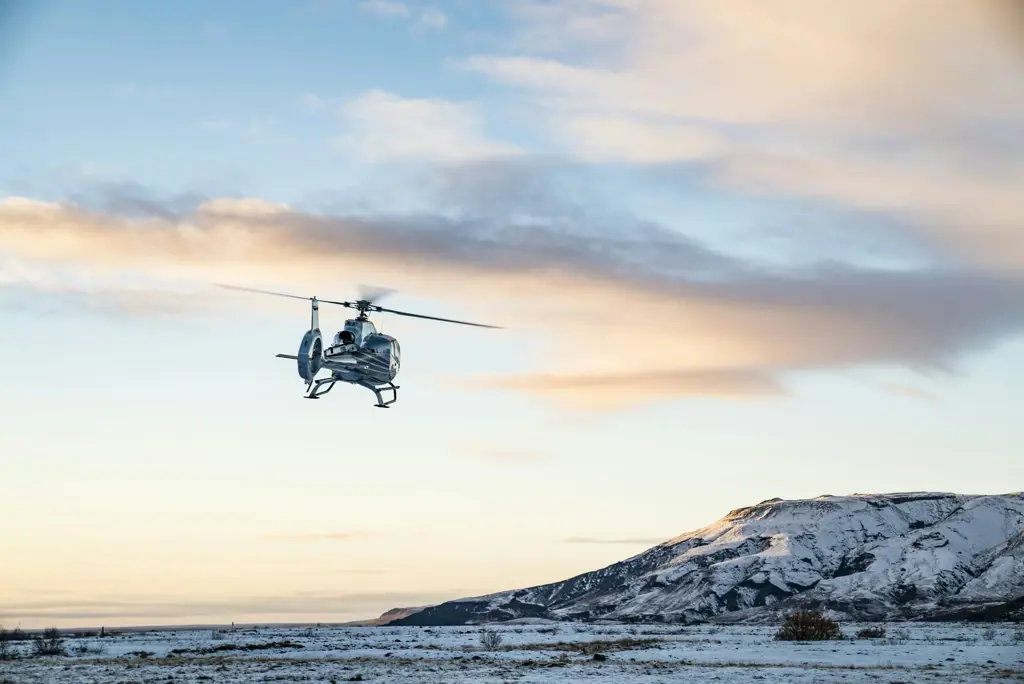 A helicopter ascends over a snow-covered landscape at sunrise, framed by pastel skies and distant mountain ridges.