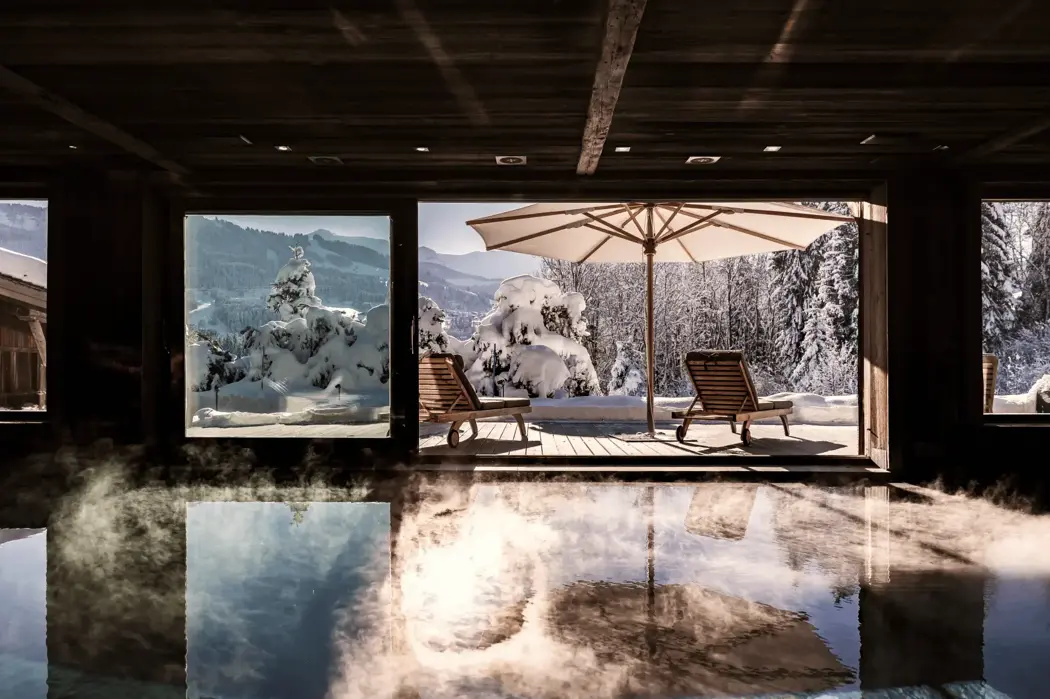 Indoor pool area with large windows overlooking a snowy mountain landscape, featuring wooden lounge chairs and a sun umbrella on the terrace outside. Steam rises gently from the warm pool water.