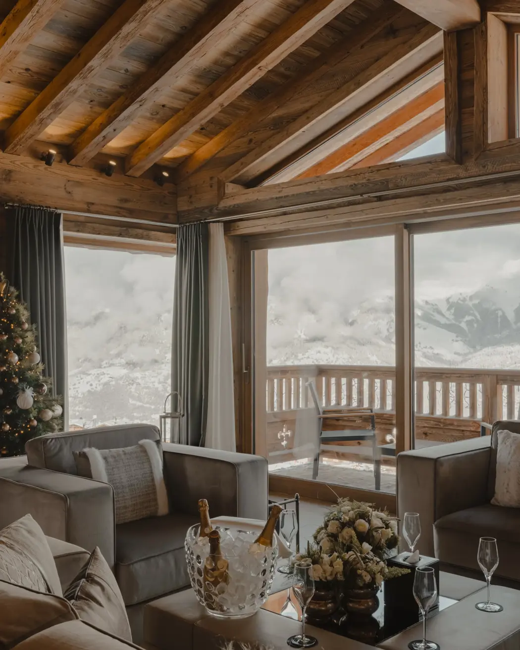 Luxury chalet living room with panoramic mountain view, wooden beams, Christmas tree, and champagne setup on the coffee table.