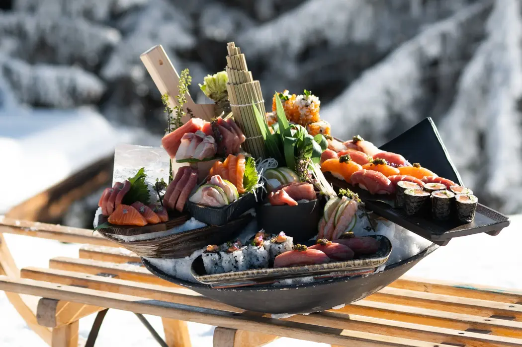 An artfully arranged sushi platter featuring fresh sashimi and rolls, displayed outdoors on a wooden sled against a snowy alpine backdrop.